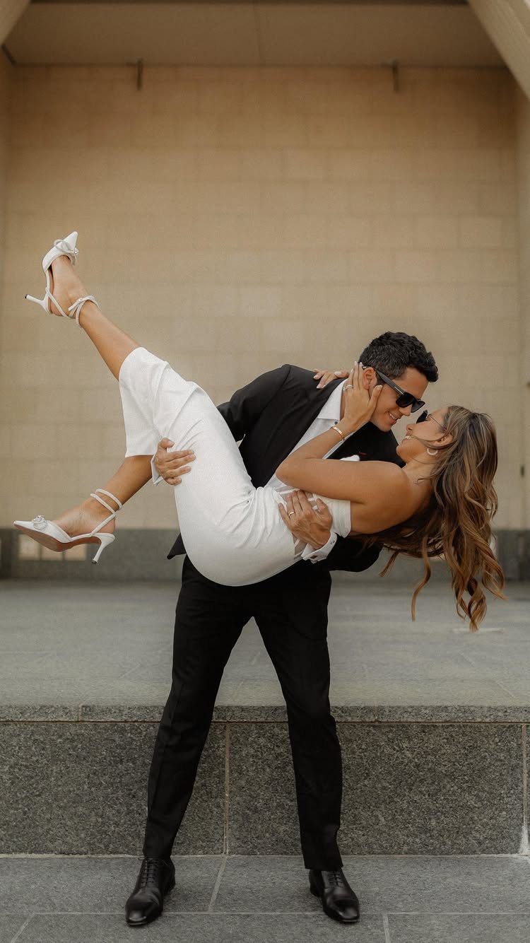 Man in black suit dipping woman in white jumpsuit against stone wall