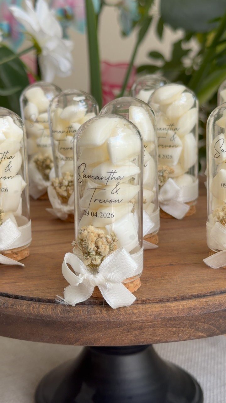 Glass vials with white petals and ribbons as wedding favors on wooden stand