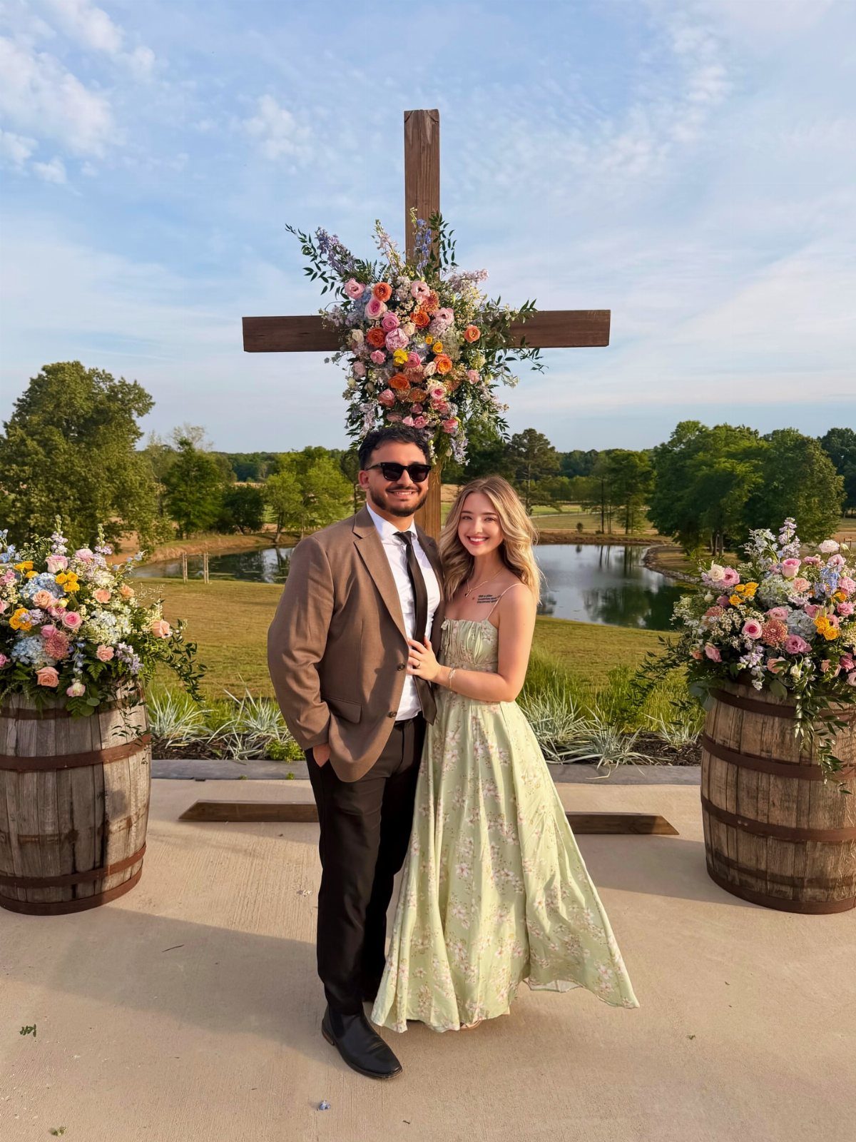 Couple posing in front of a flower-decorated wooden cross by a scenic pond.