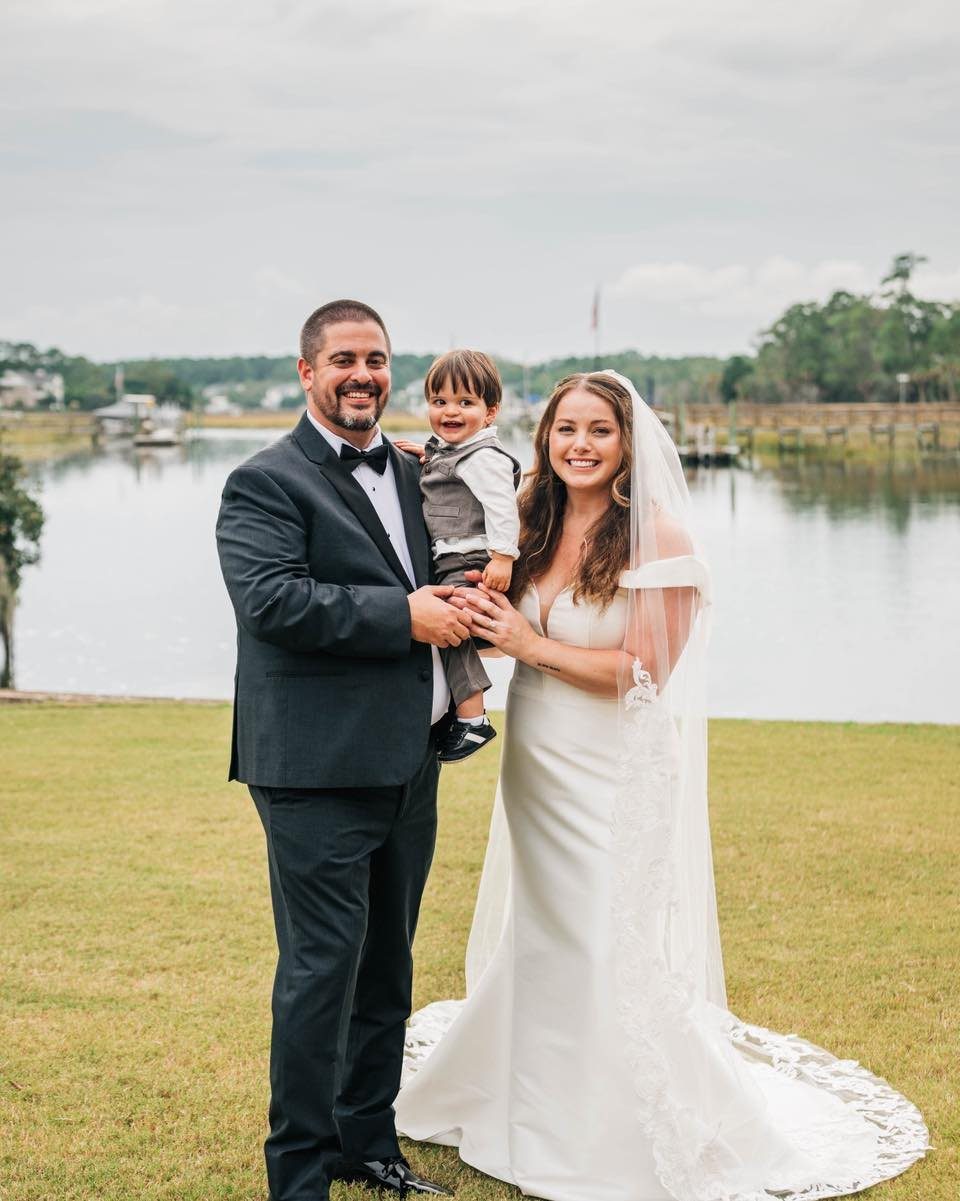 Bride and groom holding a young boy in wedding attire by a lake.