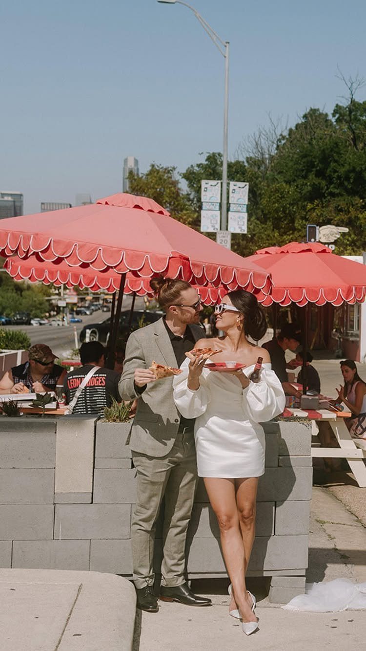 Couple in wedding attire eating pizza under red umbrellas in an outdoor setting