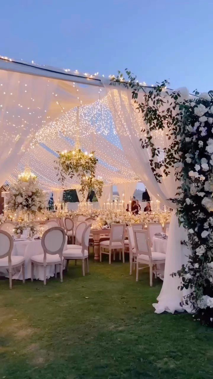 Wedding reception setup under a white tent with fairy lights and white floral decor.