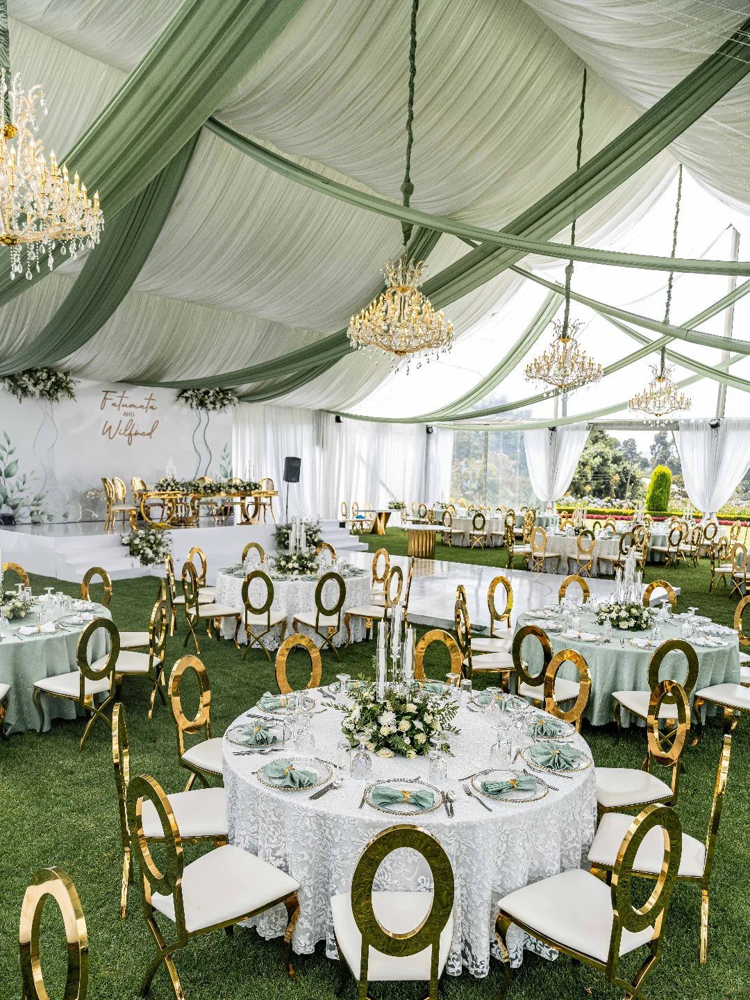 Wedding reception setup with round tables, gold chairs, and chandeliers under a draped tent.
