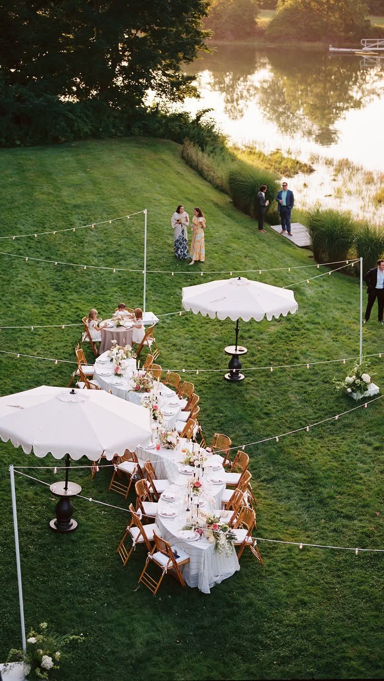 Outdoor wedding reception with long white tables, wooden chairs, and umbrellas by a lake.