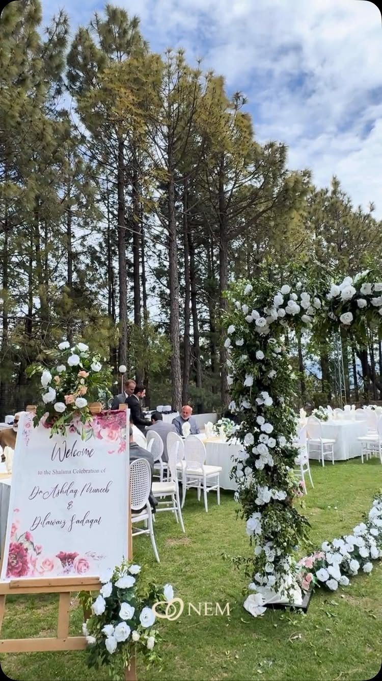 Outdoor wedding reception with white tables, chairs, and a floral arch under pine trees.