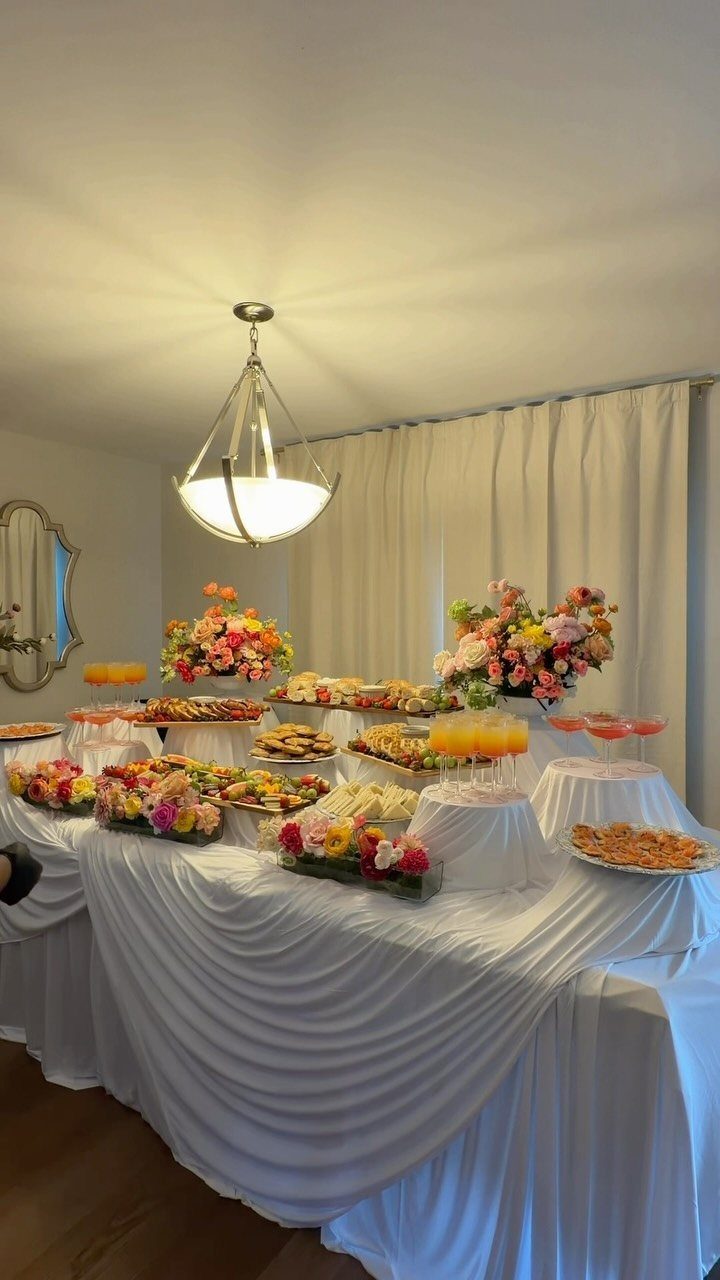 Buffet table with white pleated linens, appetizers, and colorful floral centerpieces indoors.