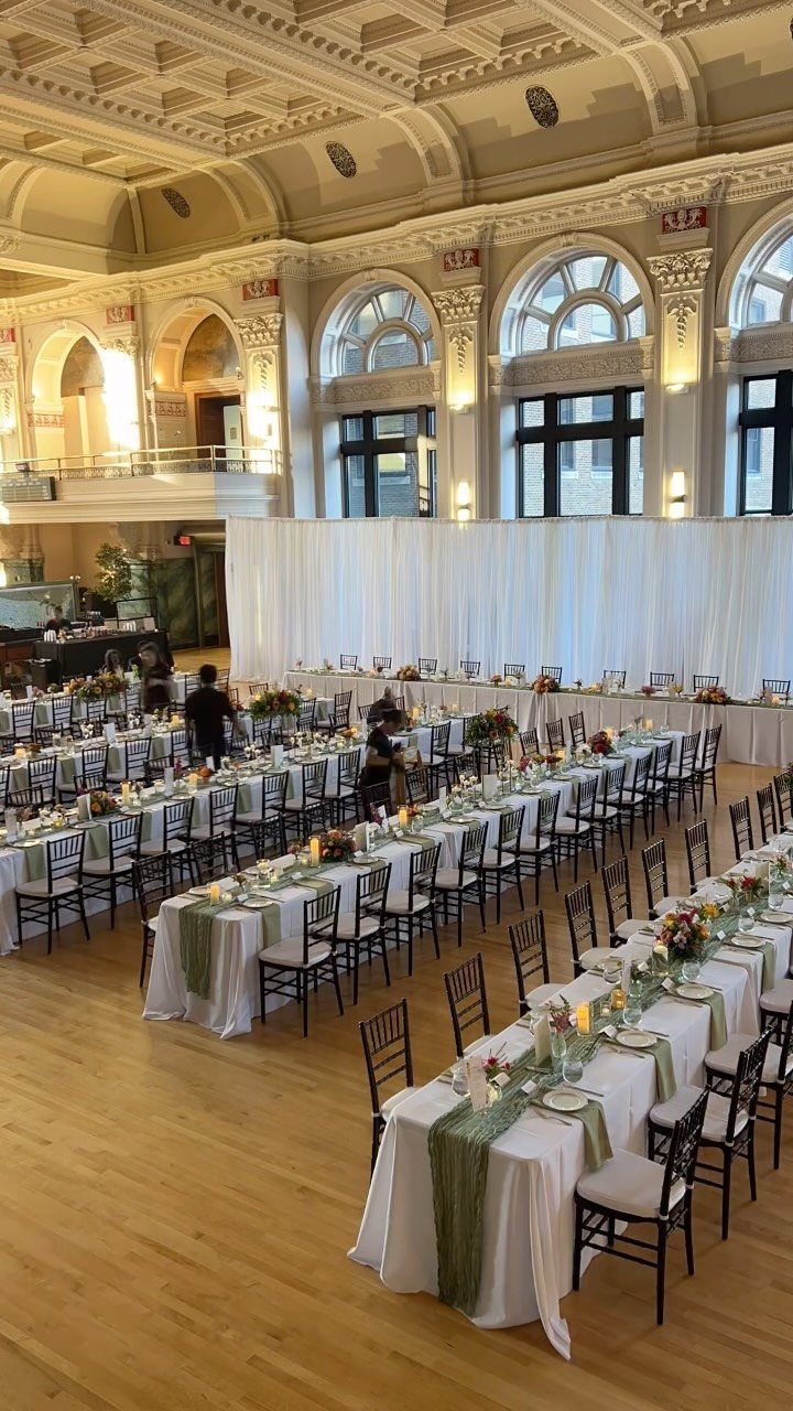 Long banquet tables with sage green runners and floral centerpieces in an ornate hall.