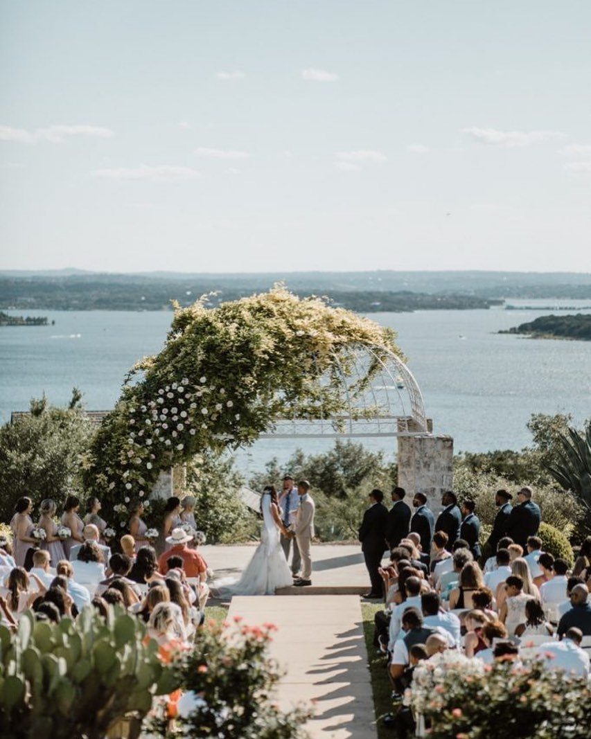 Wedding ceremony with guests seated in chairs under a floral arch by the water