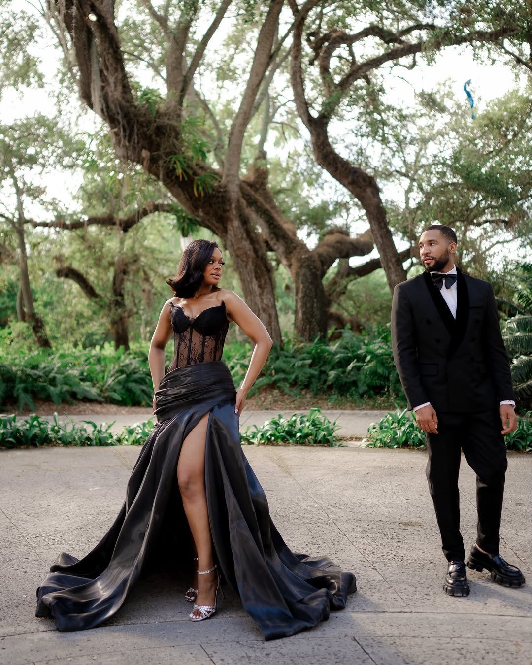 Couple in formal black wedding attire posing outdoors among green trees and foliage.