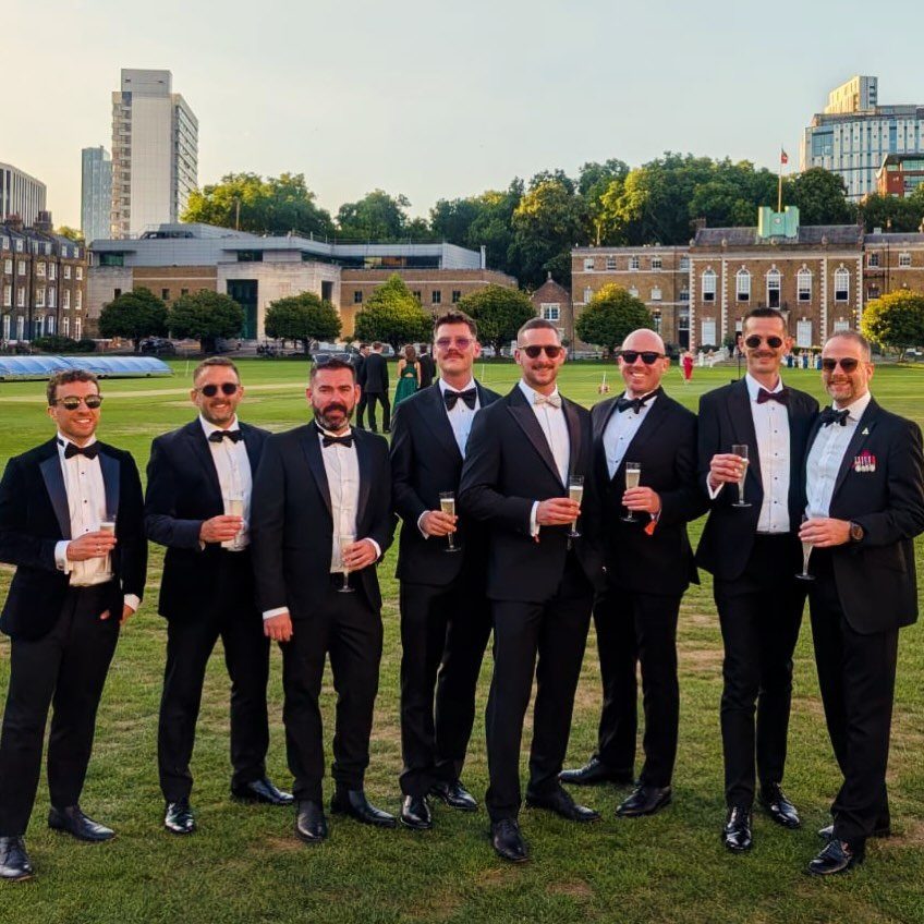 Nine men in black tuxedos and bow ties holding champagne on a lawn