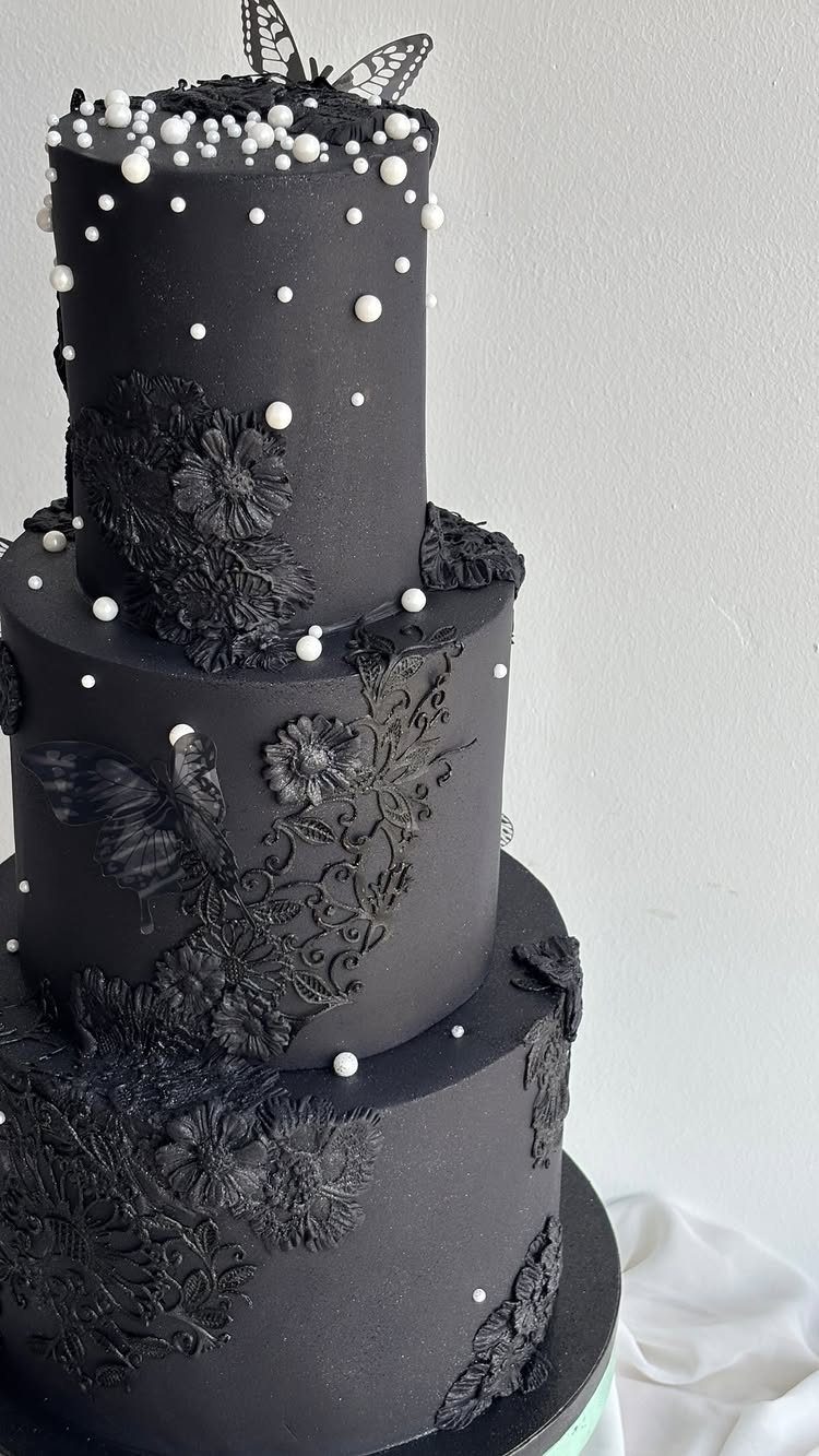 Three-tiered matte black wedding cake with floral frosting, white pearls, and butterfly topper.