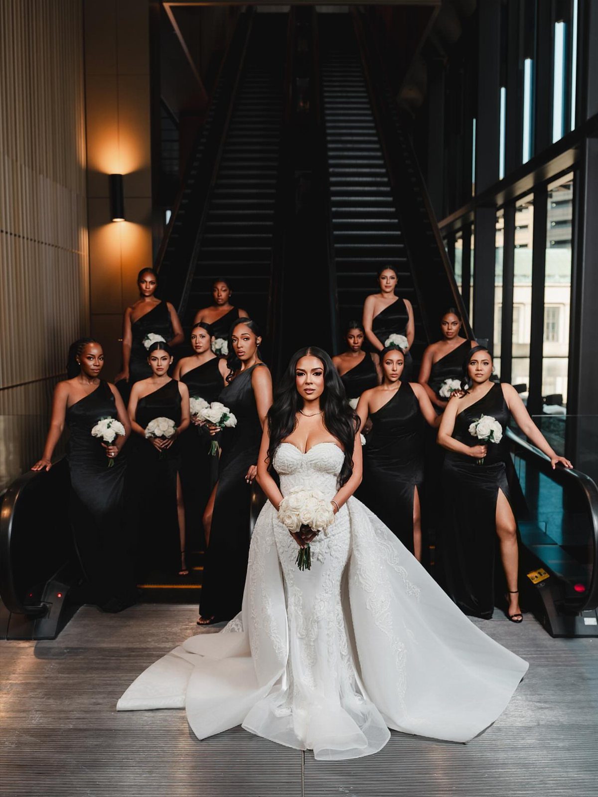 Bride in white gown surrounded by bridesmaids in black dresses on an escalator