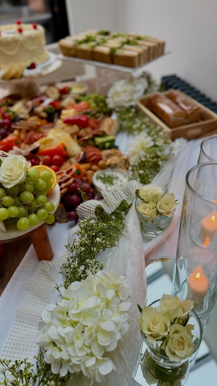 Charcuterie table with cheeses, fruits, and white floral arrangements for a party.