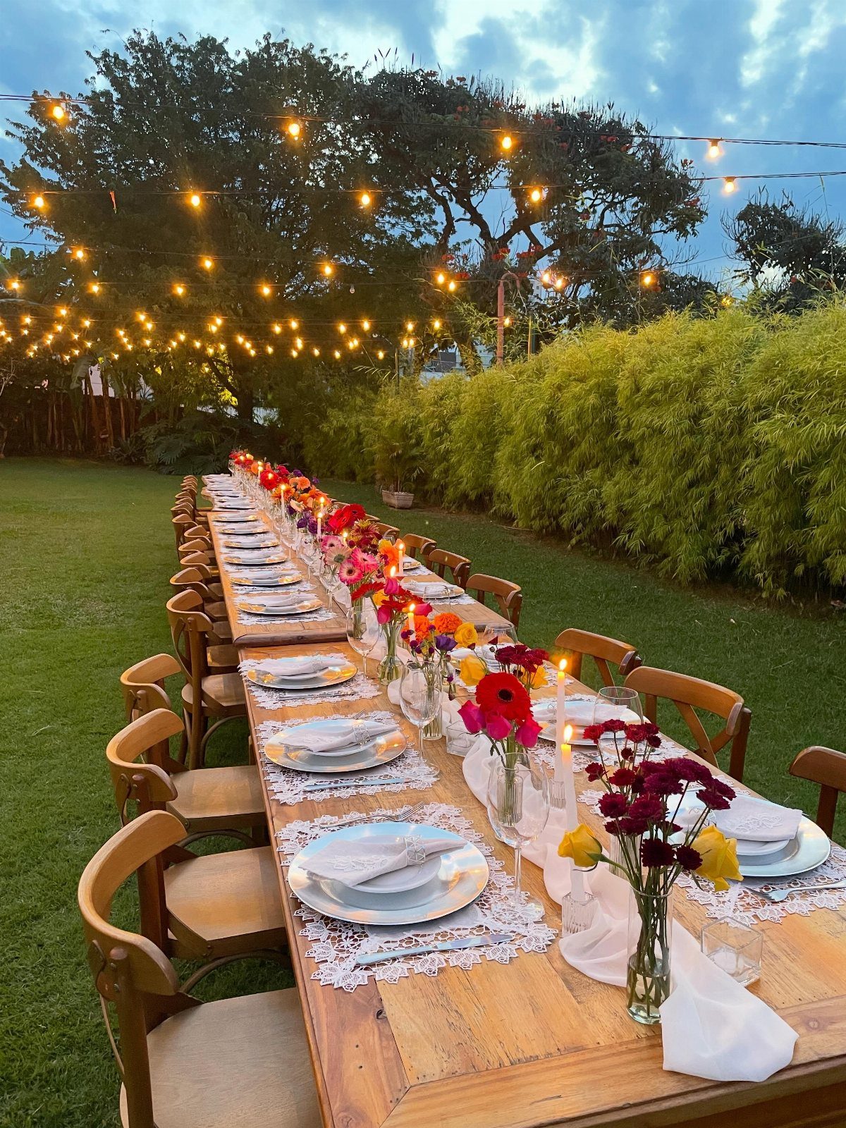 Long wooden outdoor dining table with floral centerpieces under glowing string lights at twilight.
