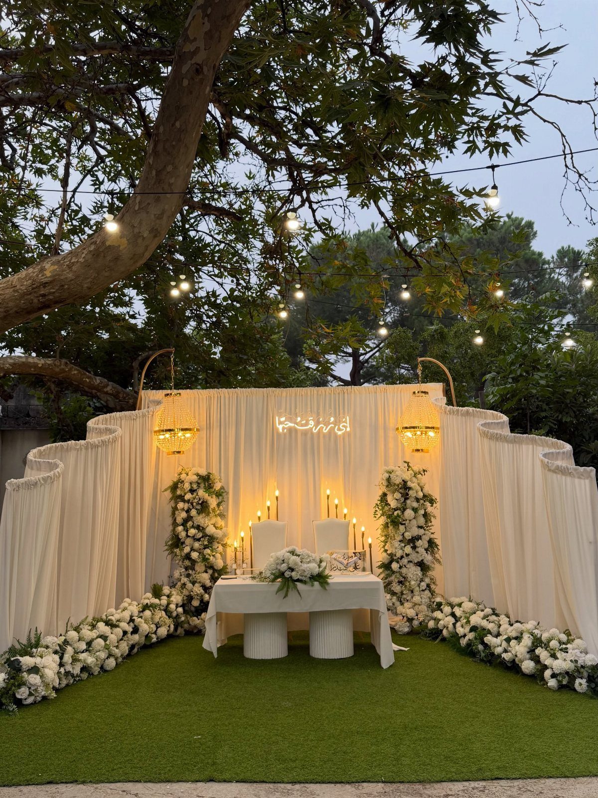 White floral arrangements and draped backdrop under a tree with string lights outdoors.