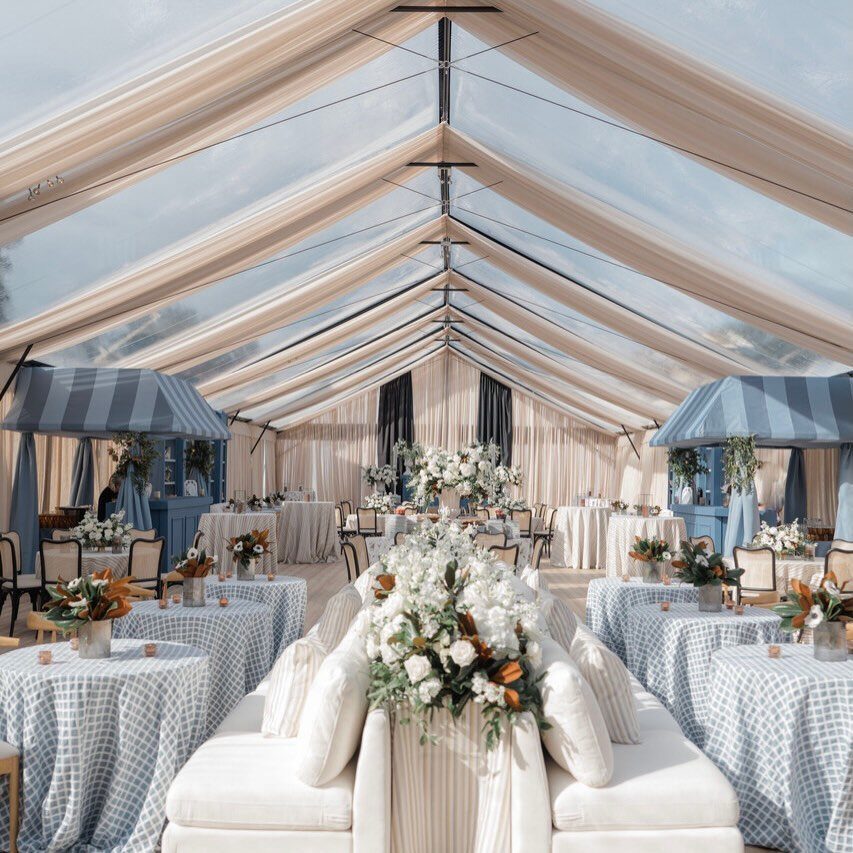 Clear-top event tent with beige ceiling draping, white sofas, and blue patterned tables.