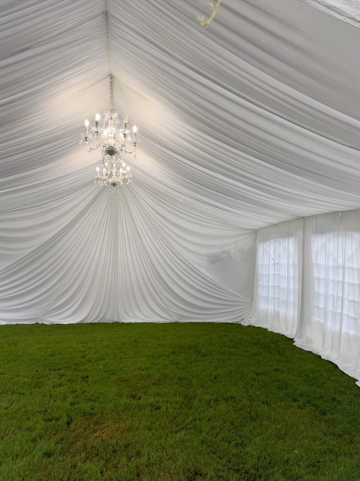 Crystal chandelier hanging inside a white draped wedding tent over green grass.