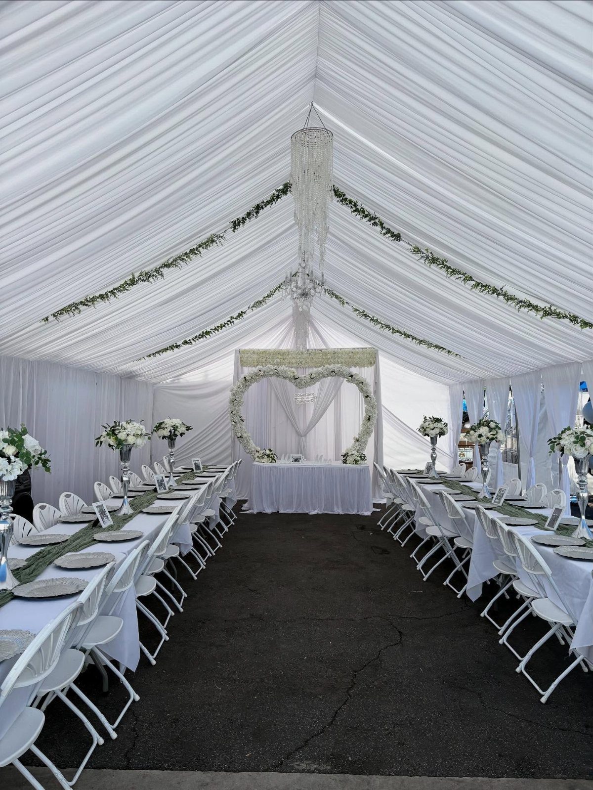 White draped wedding tent with long banquet tables and a floral heart arch.
