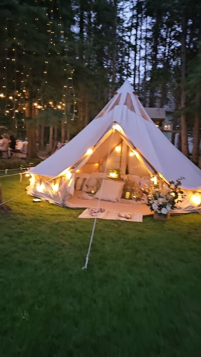 White bell tent on a lawn with warm interior lighting and surrounding trees.