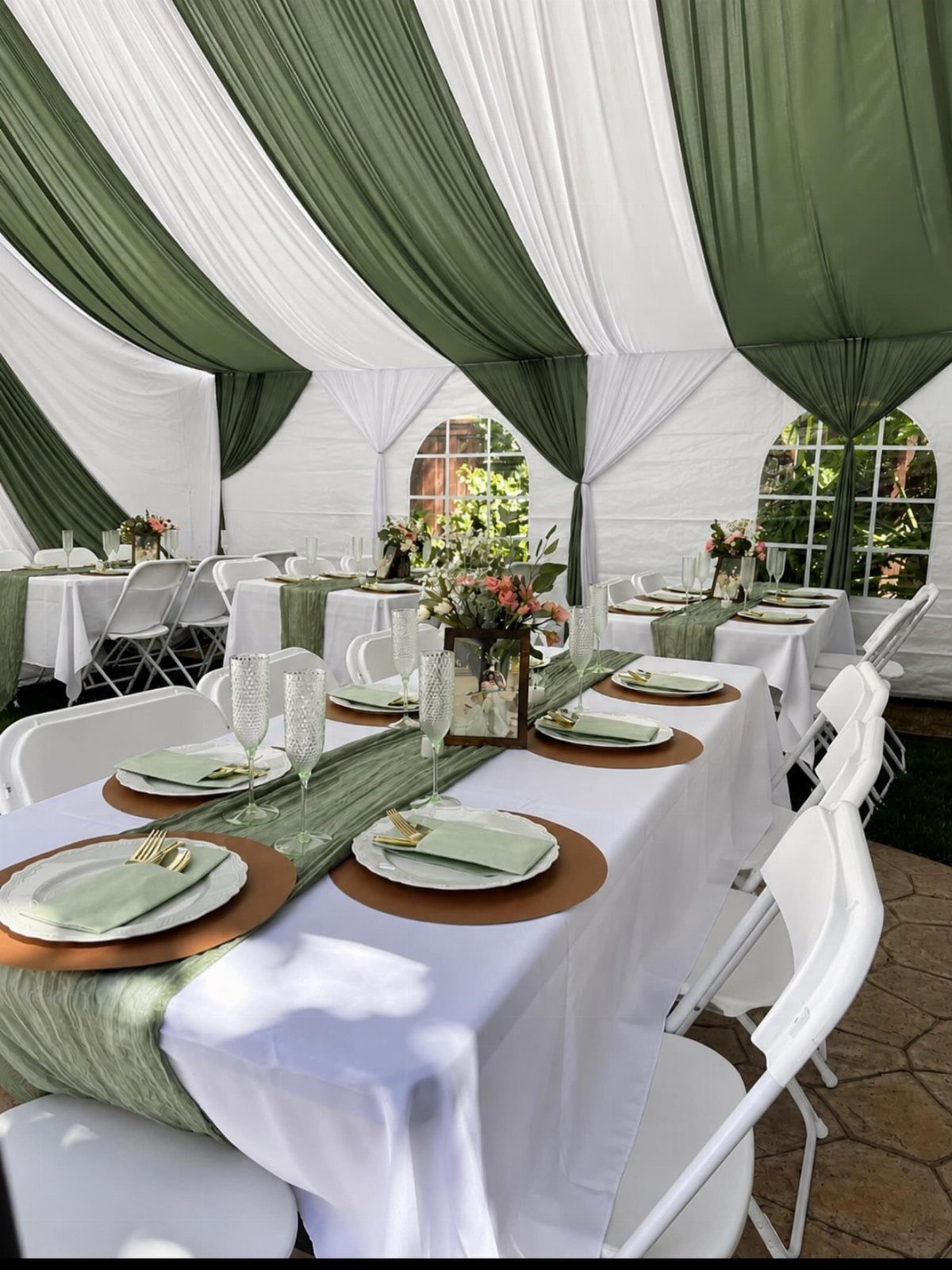 Wedding tables with white tablecloths, sage green runners, and gold chargers under a tent.