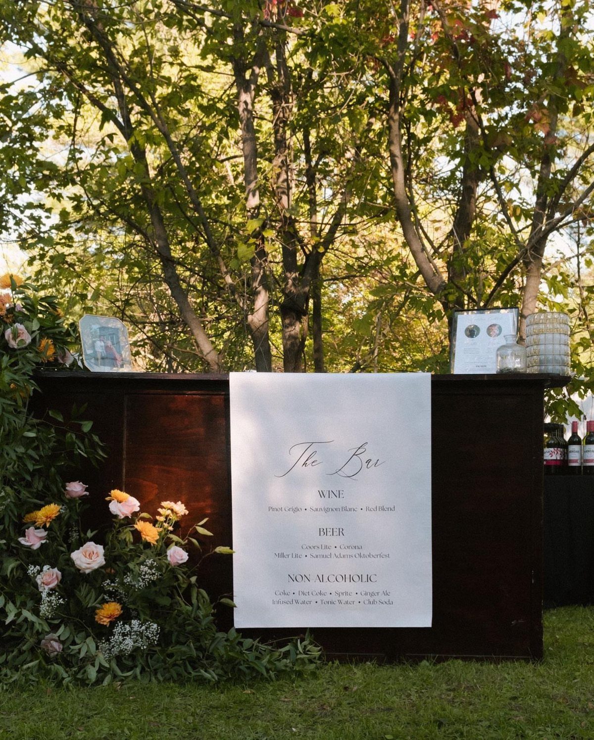Outdoor wooden bar with a drink menu and floral arrangements under green trees.