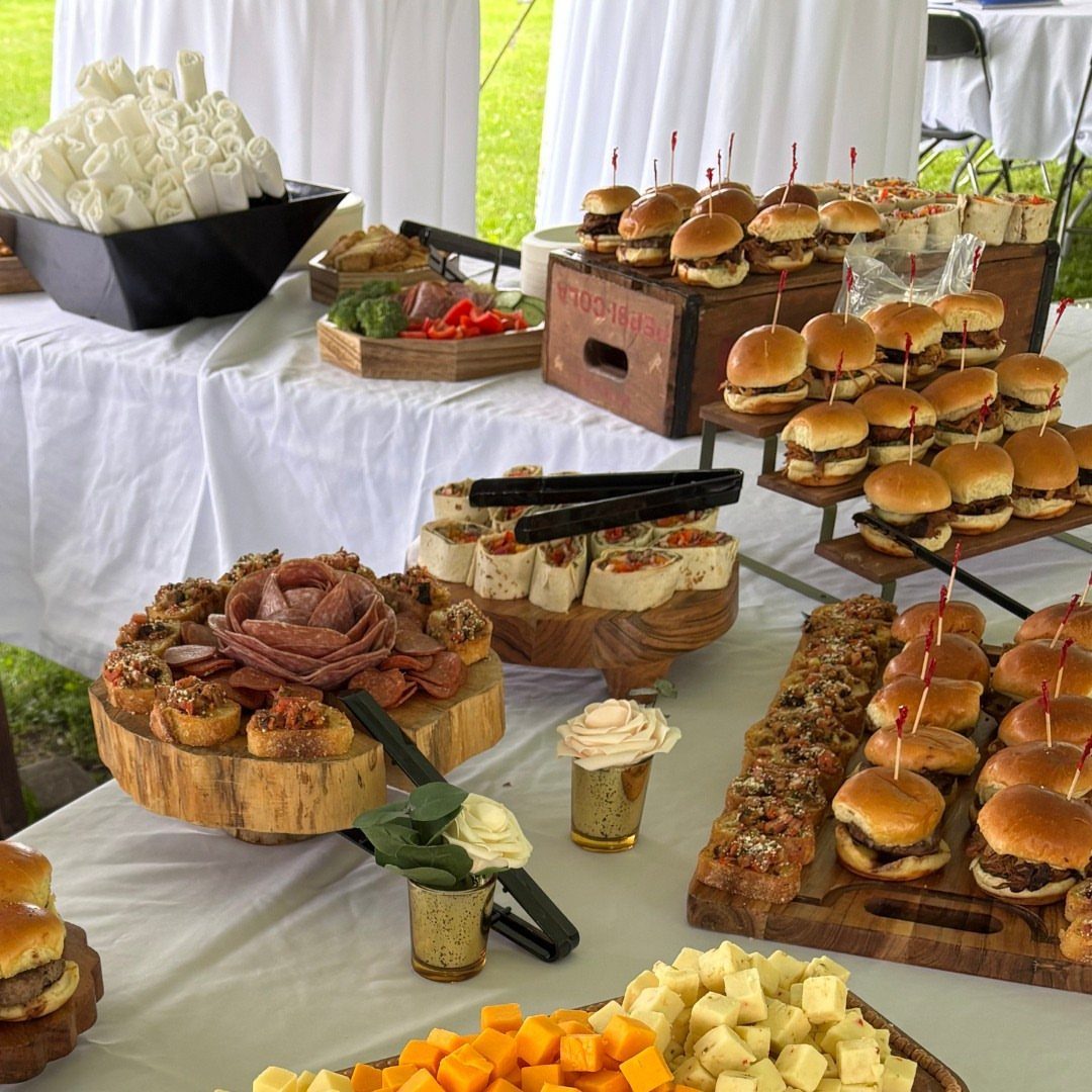 Buffet table with sliders, charcuterie, wraps, and cheese cubes on white tablecloth.