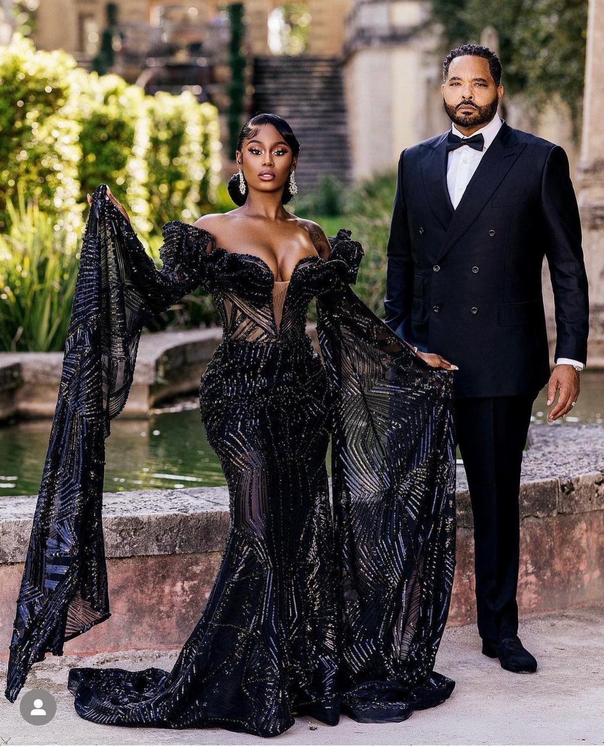 Black couple posing outdoors, woman in black shimmering gown and man in tuxedo.