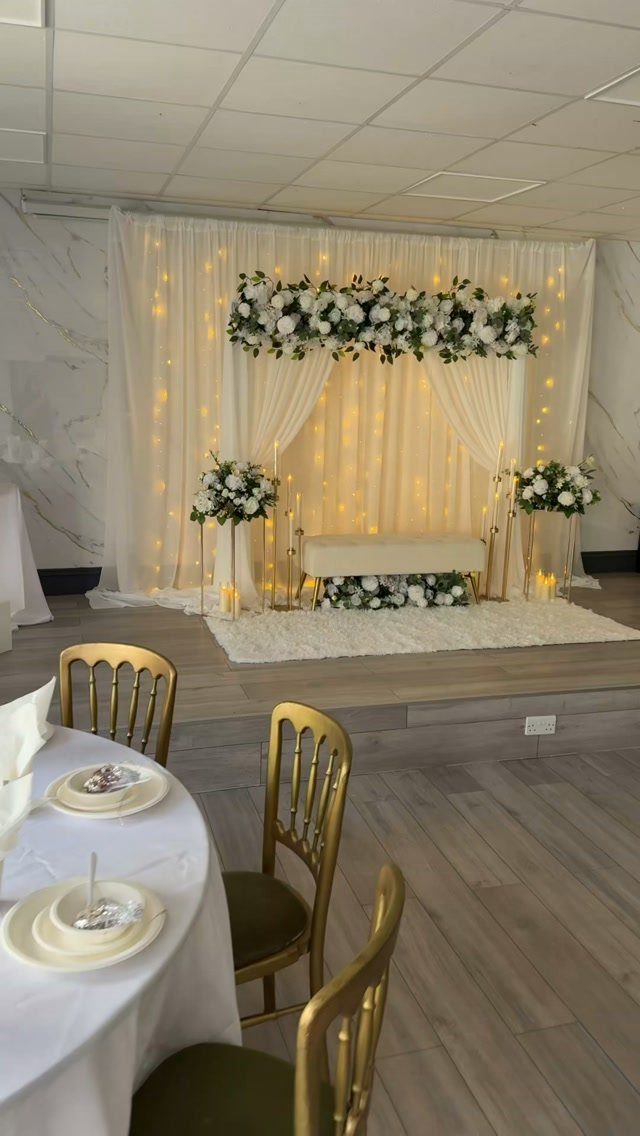 Wedding altar with white curtains, fairy lights, and white floral arrangements on gold stands.