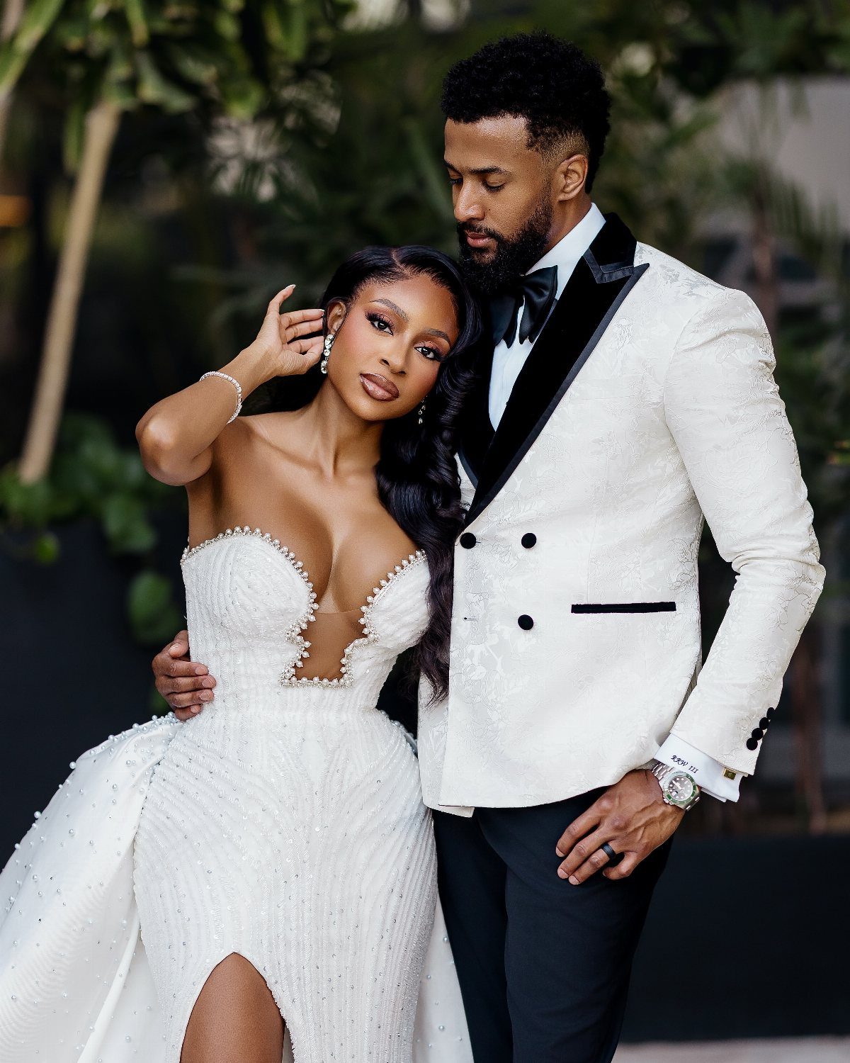 Black couple in white wedding attire posing closely together in an outdoor setting.