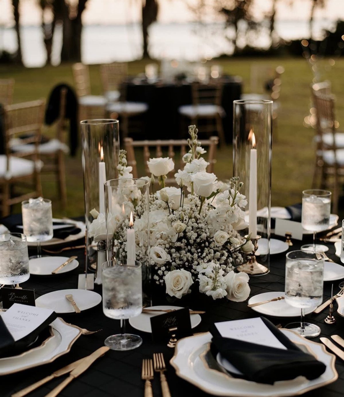 Wedding table setting with black tablecloth, white flowers, and lit candles outdoors.