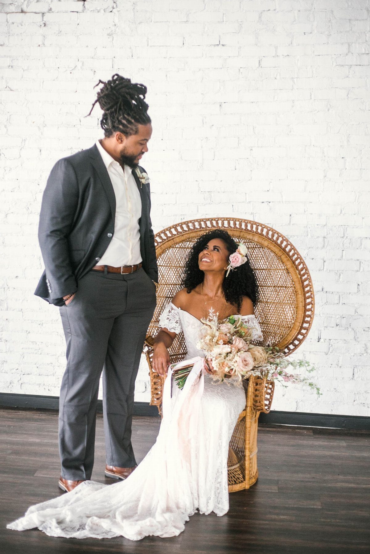 Black bride in white lace off-shoulder gown sitting in a wicker peacock chair.