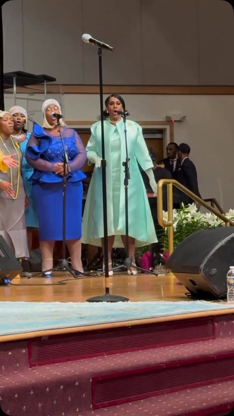 Black women singing in a choir on a stage wearing colorful formal dresses