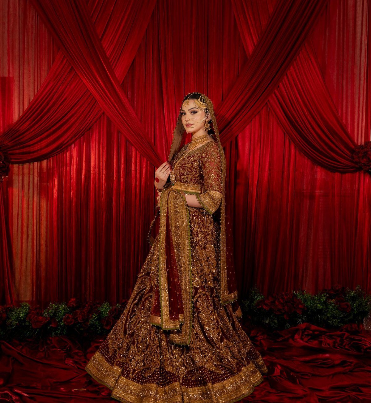Bride in gold embroidered lehenga wearing a maang tikka against red curtains