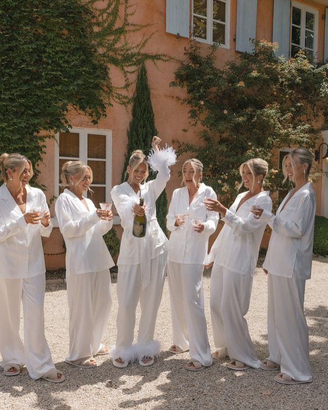 Six women in white pajamas celebrating with champagne outside a peach-colored villa.