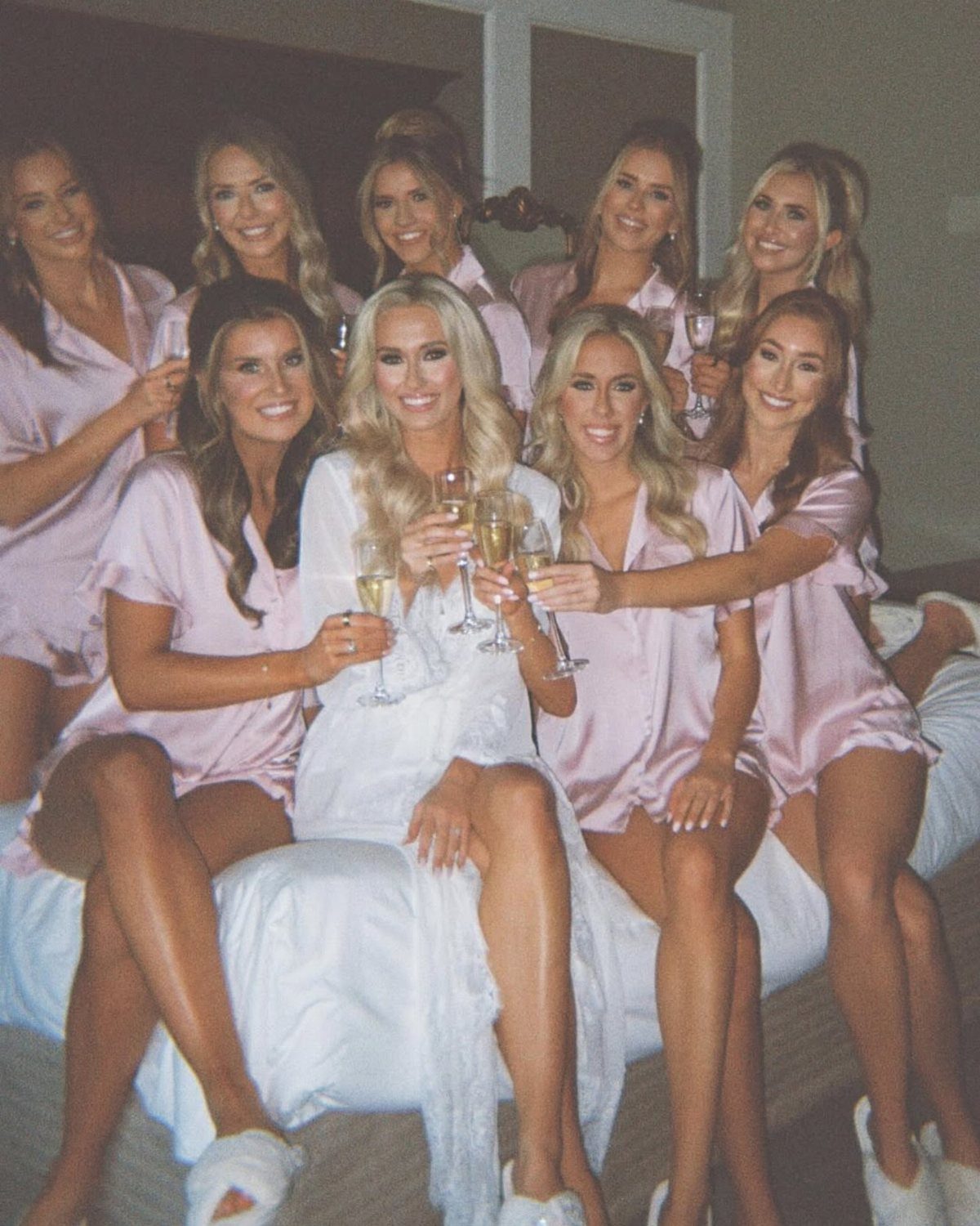 Bride in white robe and bridesmaids in pink robes holding champagne glasses on bed