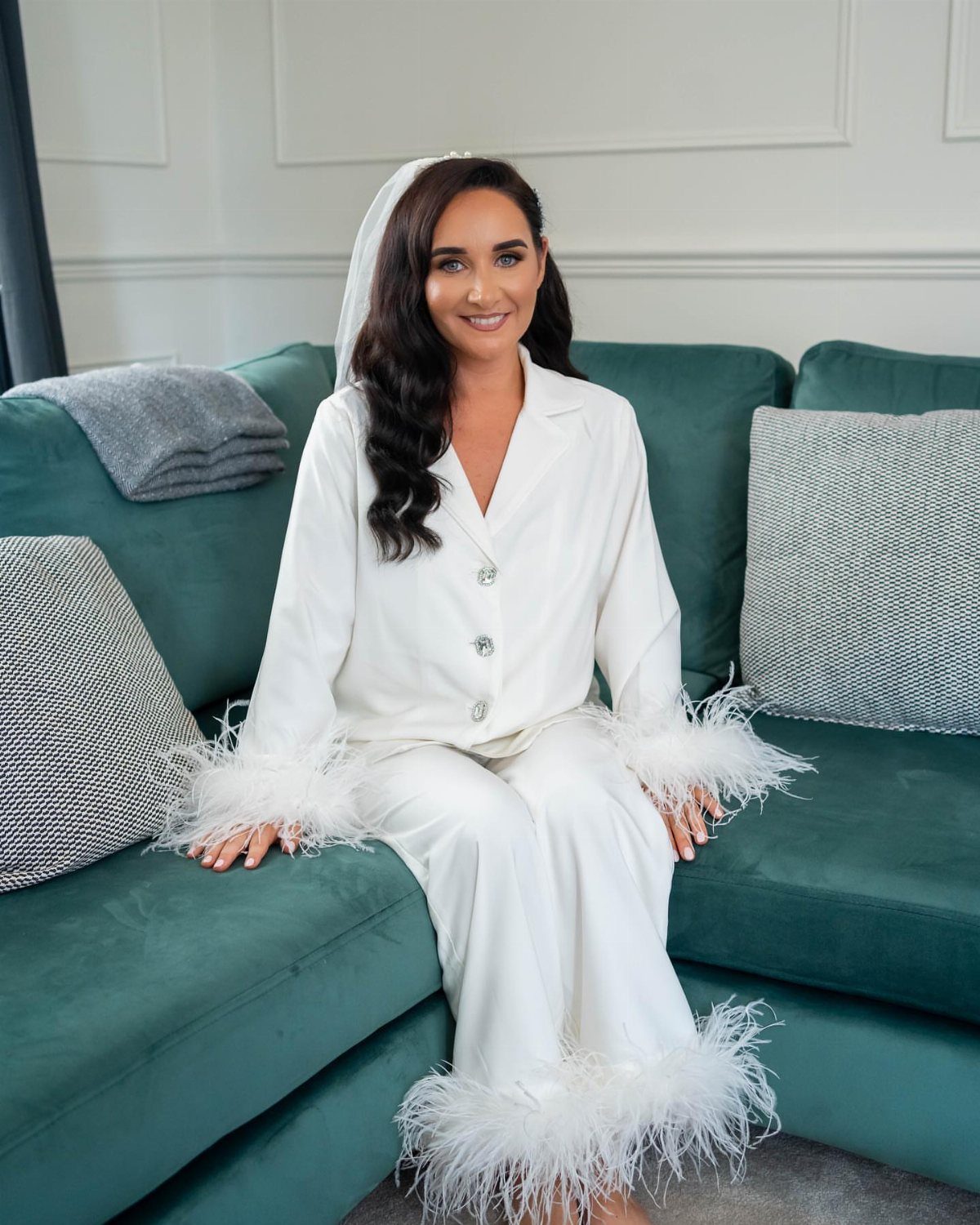Woman in white satin pajamas with feather trim sitting on a teal sofa