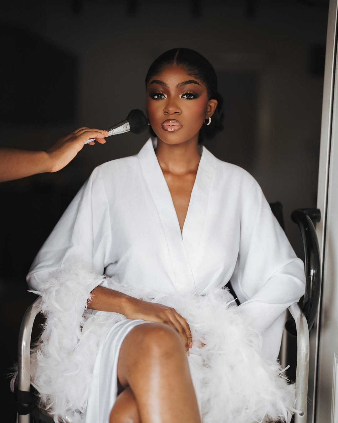 Woman in white feathered robe receiving makeup application before her wedding day