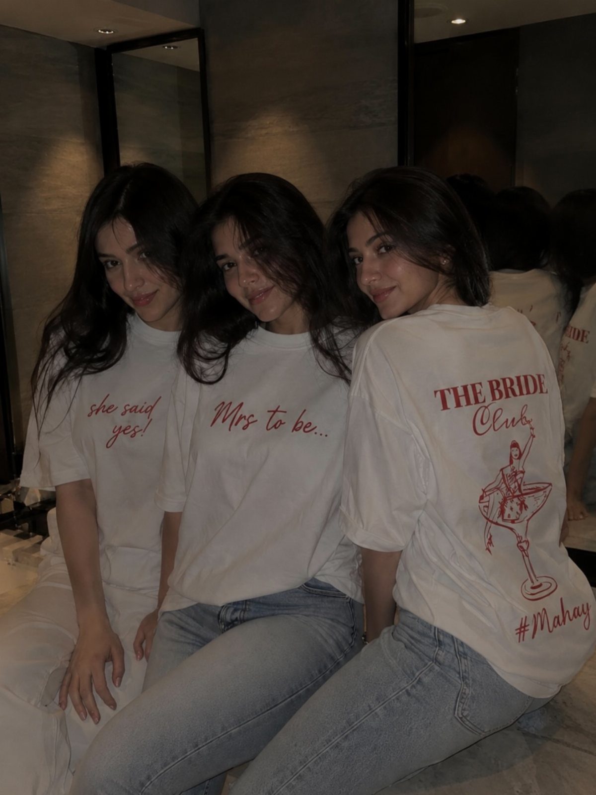 Three women wearing oversized white bridal t-shirts and jeans sitting together indoors.