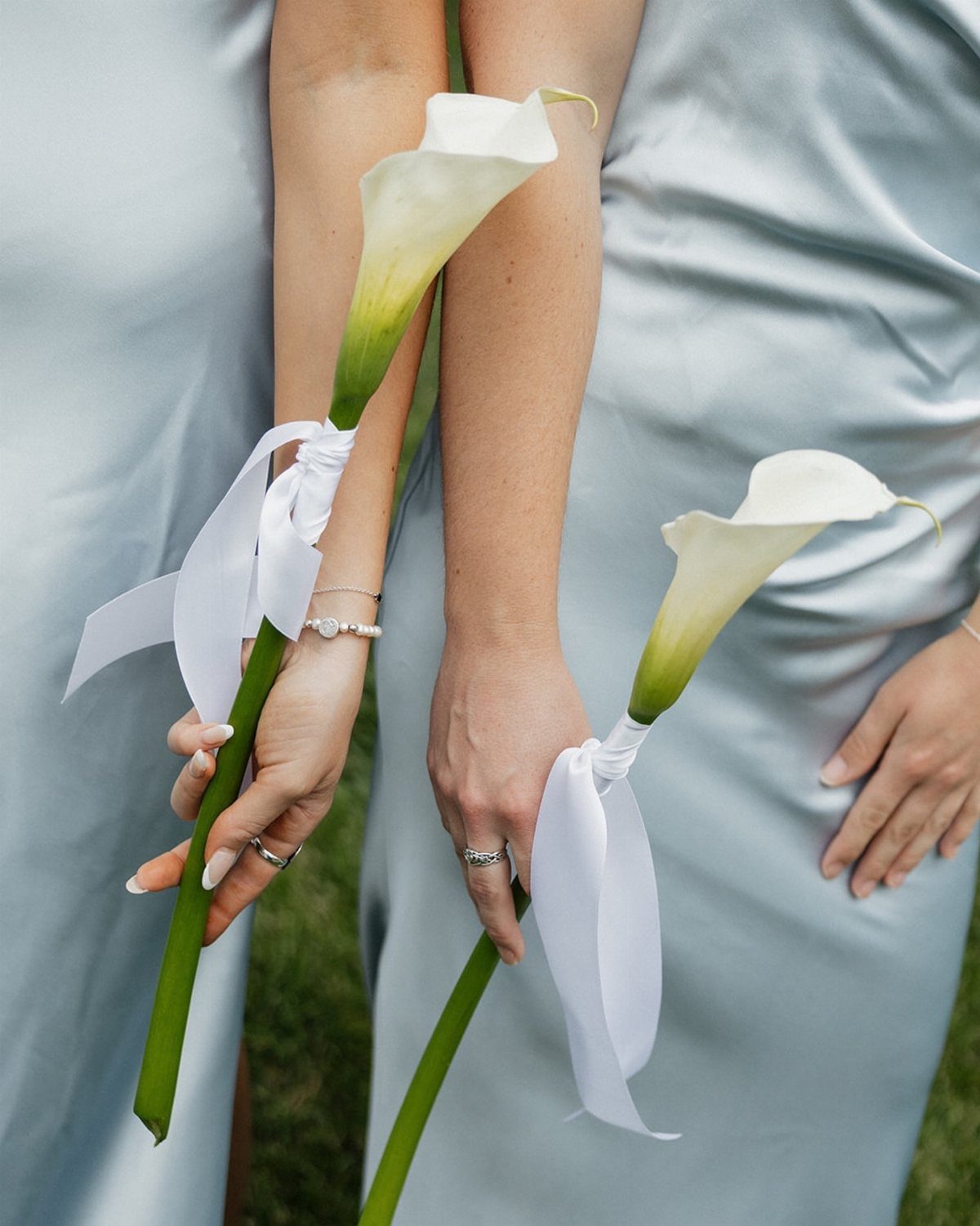 Two bridesmaids in blue dresses holding single white calla lilies with white ribbons.