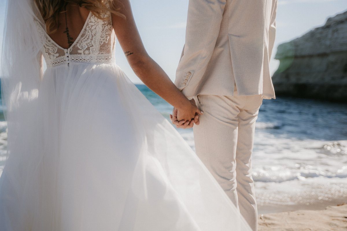 Bride and groom holding hands on a beach, groom wearing a beige linen suit.