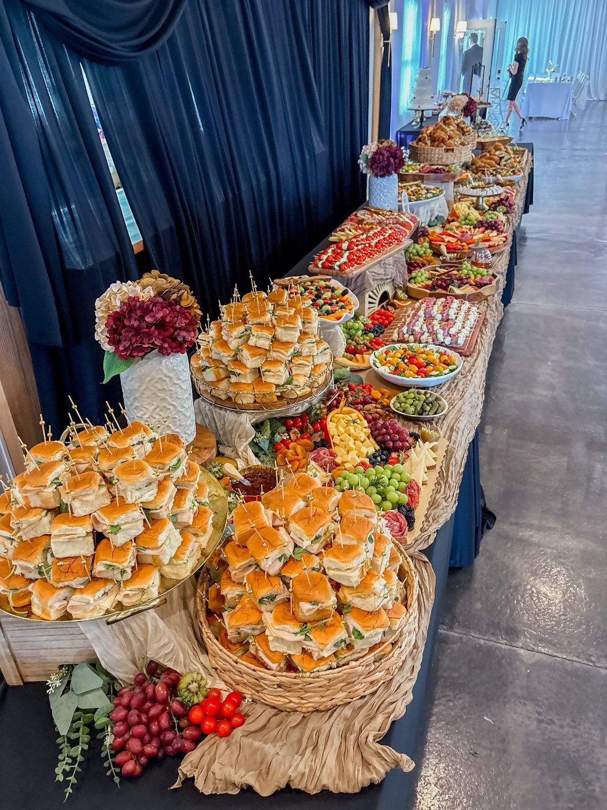 Buffet table with sliders, fruit, and appetizers set against dark blue curtains.