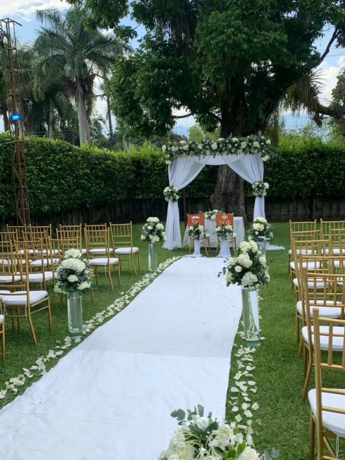 White floral arrangements in glass vases lining a white wedding aisle outdoors.