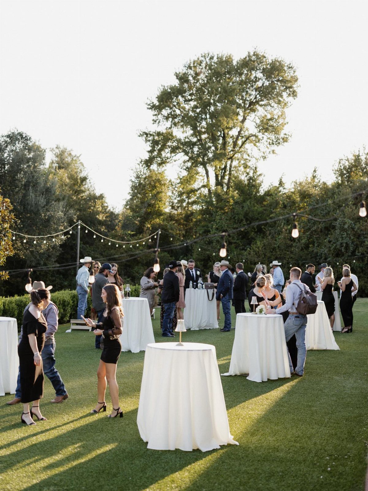 Guests mingling around white cocktail tables on a lawn with overhead string lights.