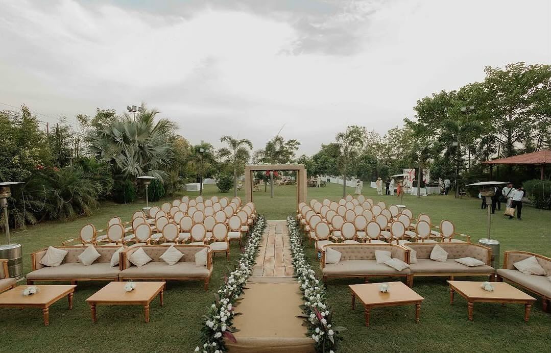 Outdoor wedding ceremony setup with beige couches, wooden tables, and rows of chairs.