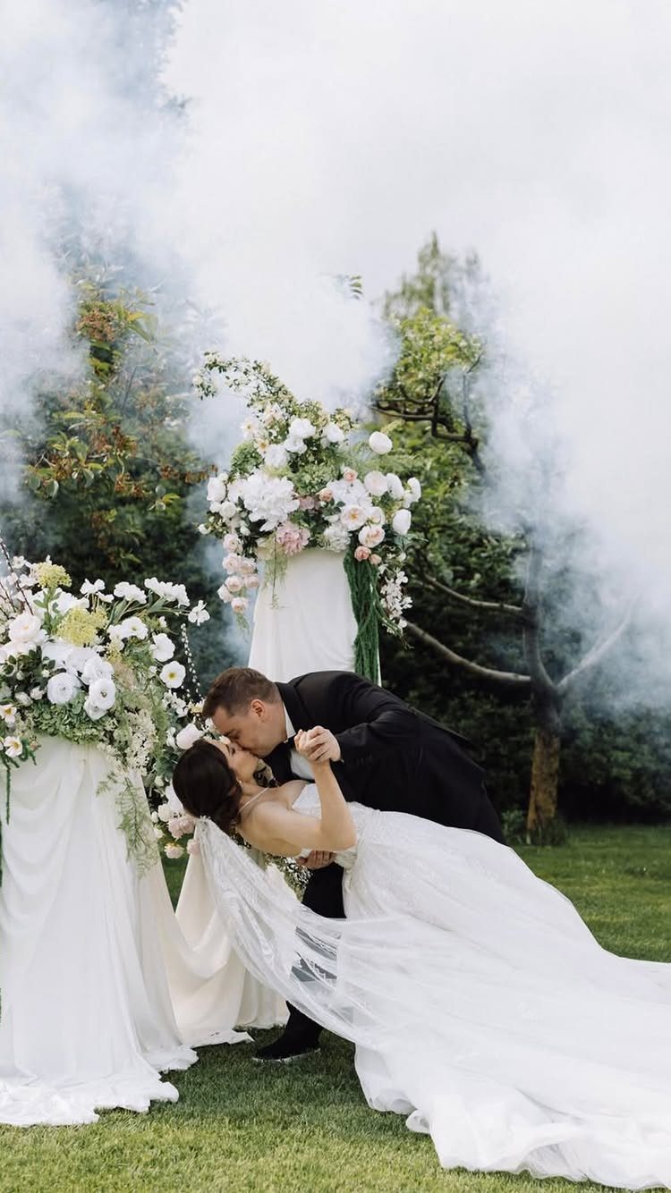 Bride and groom dip-kissing on a lawn surrounded by white smoke and flowers.