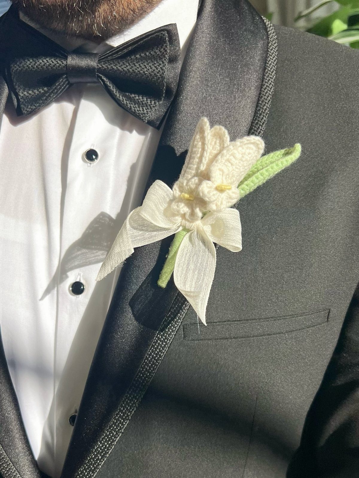 White crocheted flower boutonniere with a ribbon pinned to a black tuxedo lapel