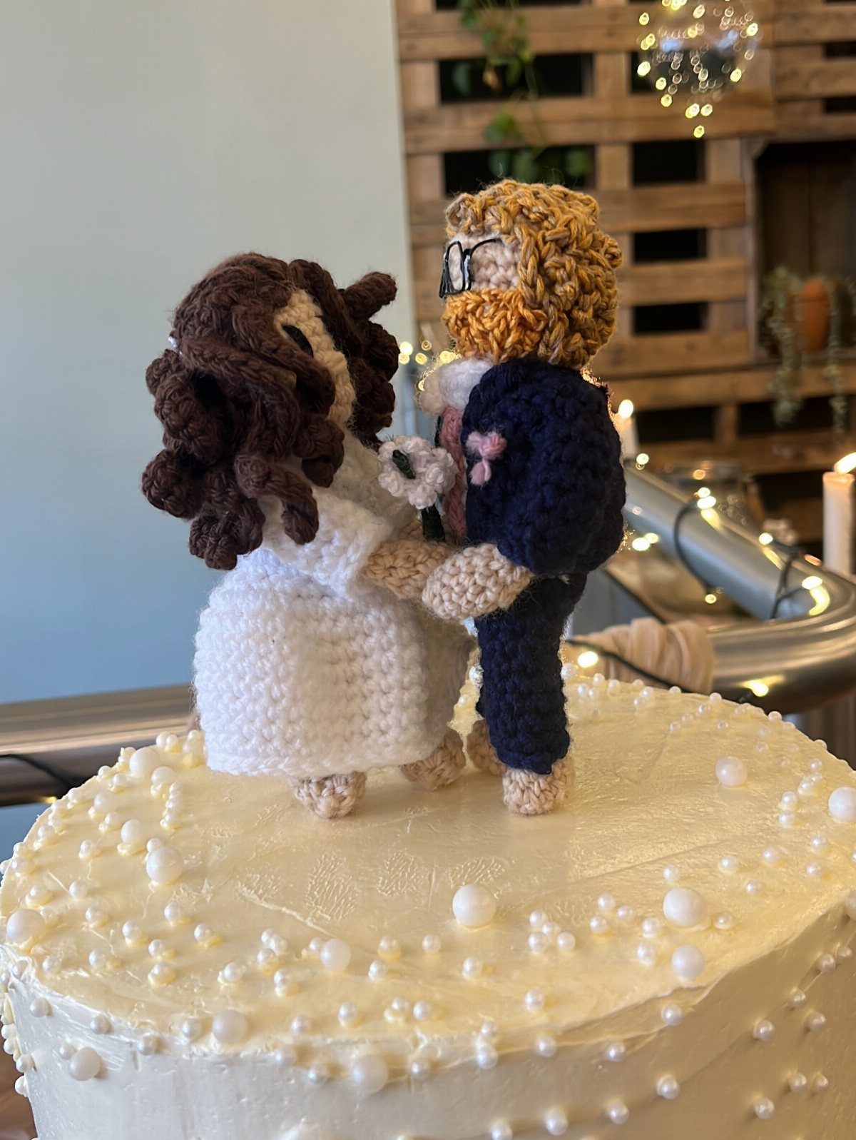 Crochet bride and groom cake toppers standing on a white pearl-decorated cake
