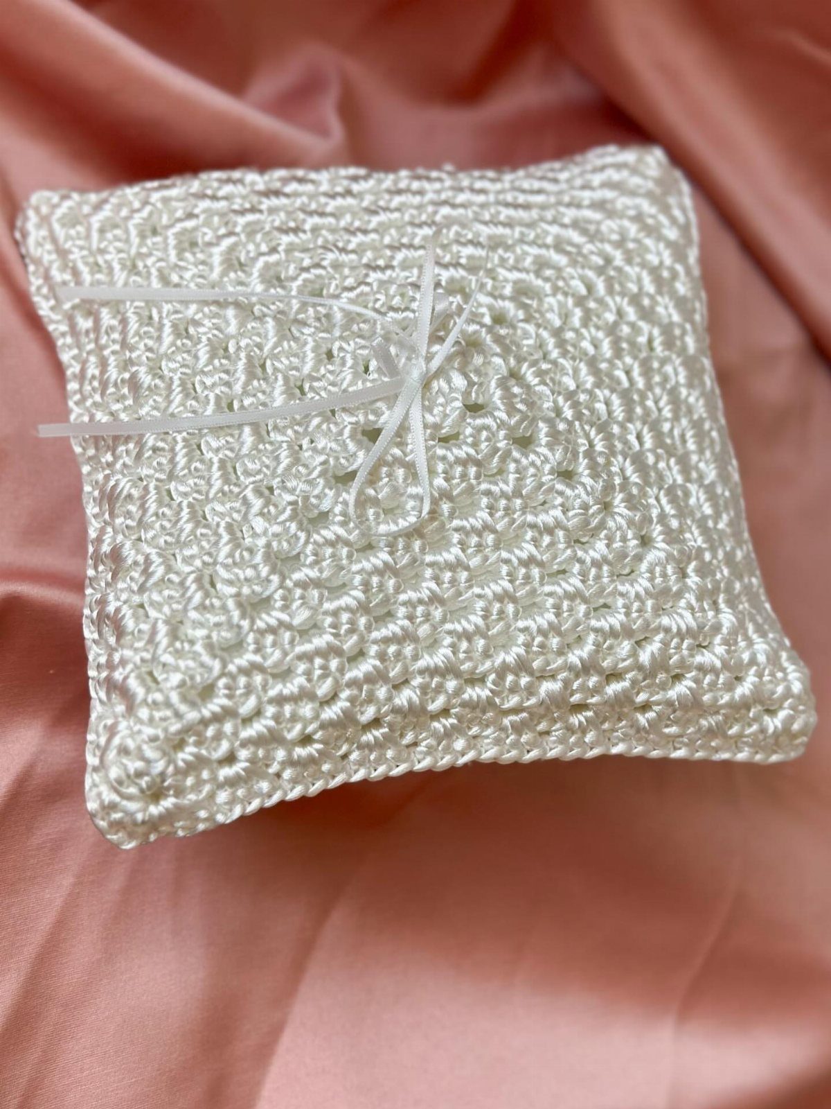 White crochet ring bearer pillow with a ribbon bow on pink satin fabric