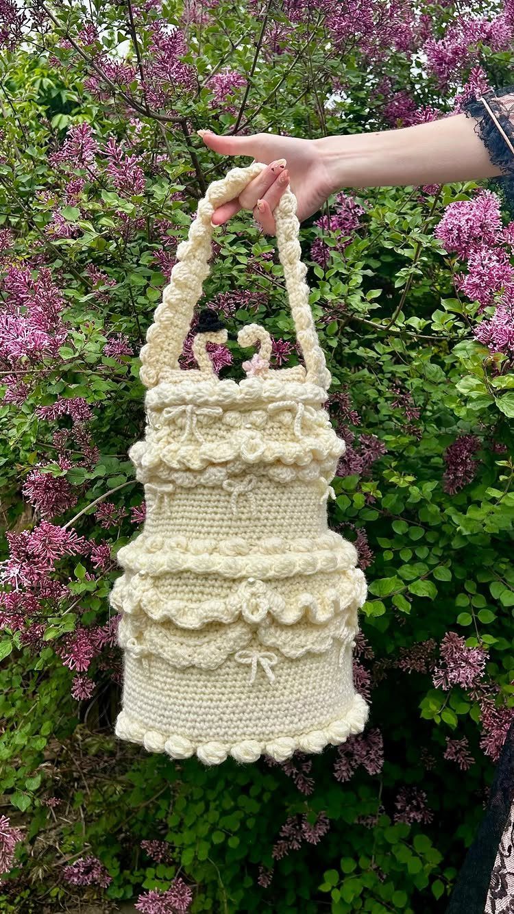 Cream crochet handbag shaped like a tiered wedding cake held against lilac bushes.