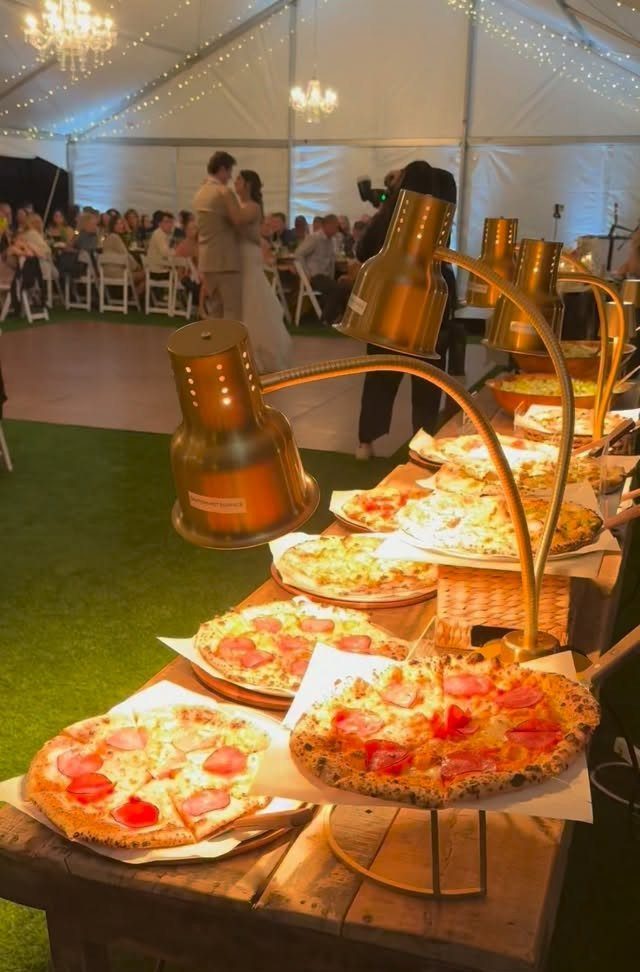 Pepperoni pizzas under gold heat lamps at a wedding reception in a tent.